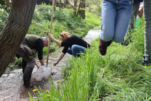 Zwei Erwachsene arbeiten an einem Bach, während ein Kind im Vordergrund springt.