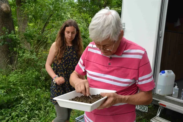 Ein älterer Mann untersucht in einer weißen Schale Erde oder Kompost, während eine junge Frau daneben im Grünen steht.