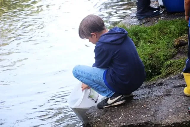 Ein Junge hockt am Ufer eines Gewässers und hält einen weißen Eimer ins Wasser.