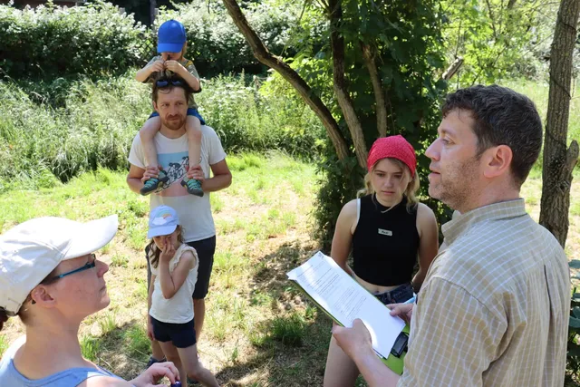 Mehrere Erwachsene und Kinder stehen bei sonnigem Wetter in einer grünen, bewaldeten Umgebung zusammen; eine Person hält ein Blatt Papier und scheint zu sprechen.
