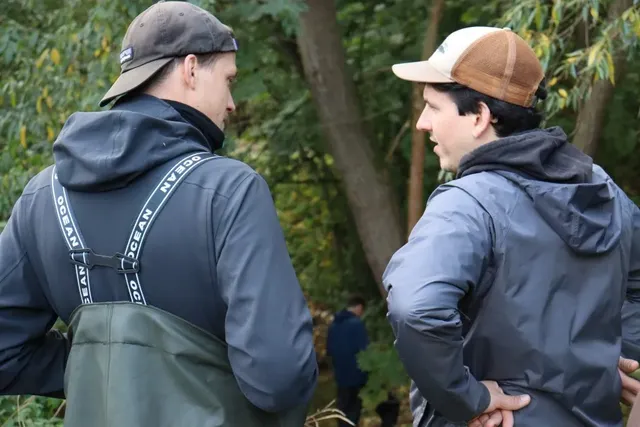 Zwei Männer sprechen draußen im Grünen miteinander, beide tragen Outdoor-Kleidung und Caps.