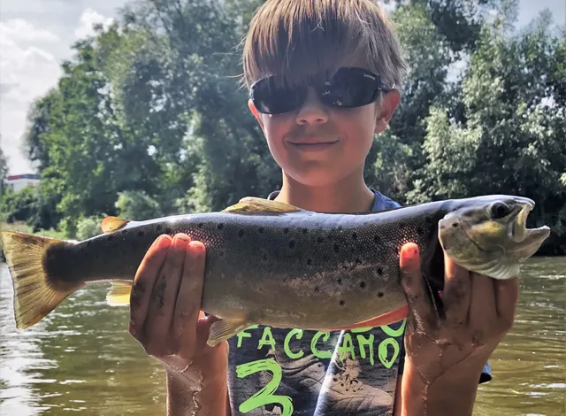 Ein Junge mit Sonnenbrille hält stolz einen großen Fisch vor einem Flussufer.