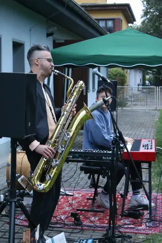 Zwei Musiker spielen draußen; einer spielt Saxophon, der andere Keyboard. Sie stehen unter einem grünen Pavillon.
