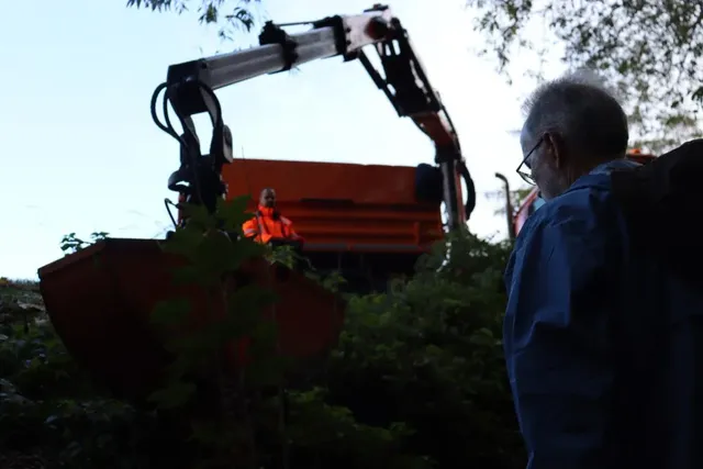 Ein Mann beobachtet einen Bagger mit Arbeiter im Hintergrund, umgeben von Büschen und Bäumen.