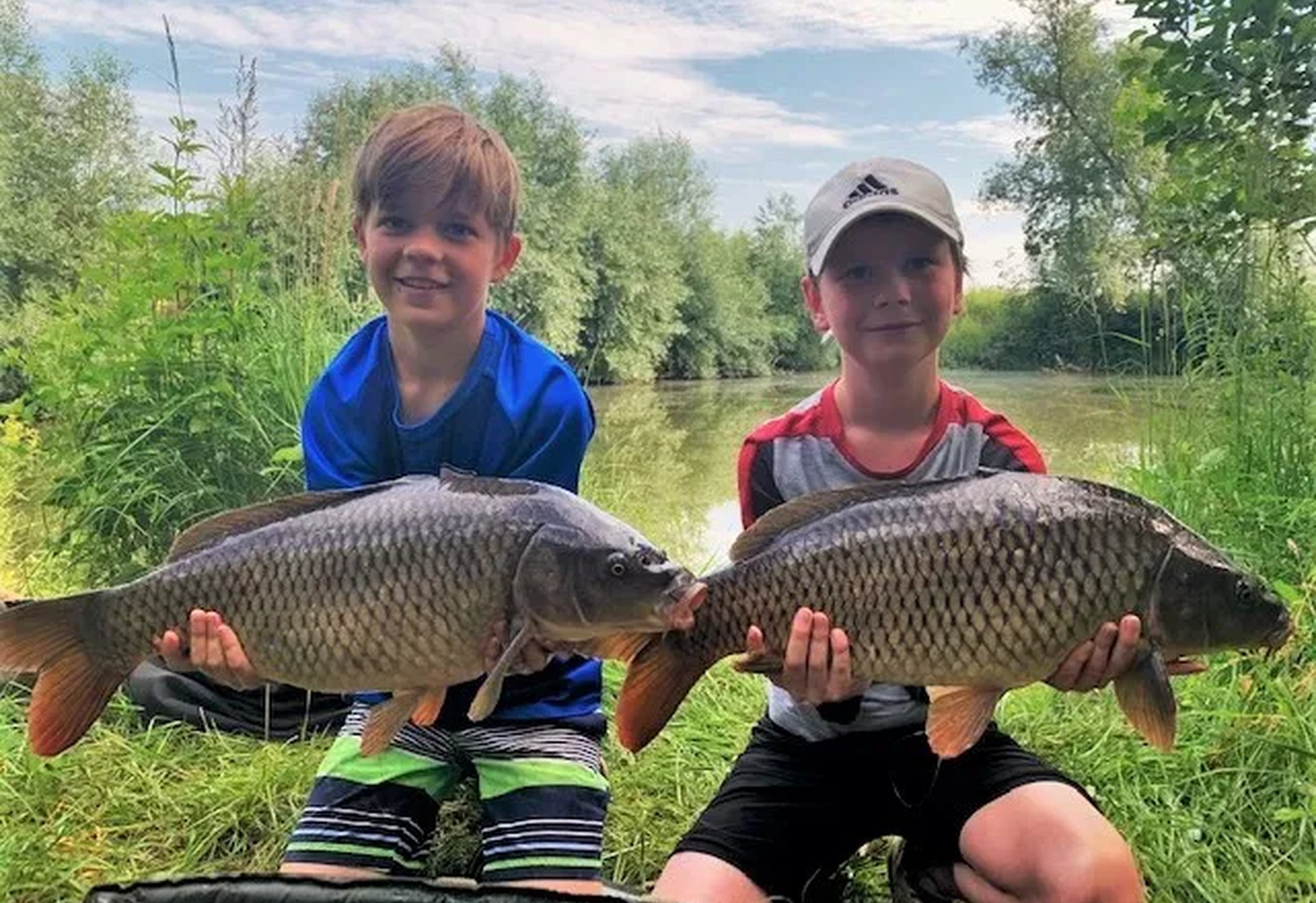 Zwei Jungen sitzen am Ufer eines Sees und halten jeweils einen großen gefangenen Fisch in den Händen.