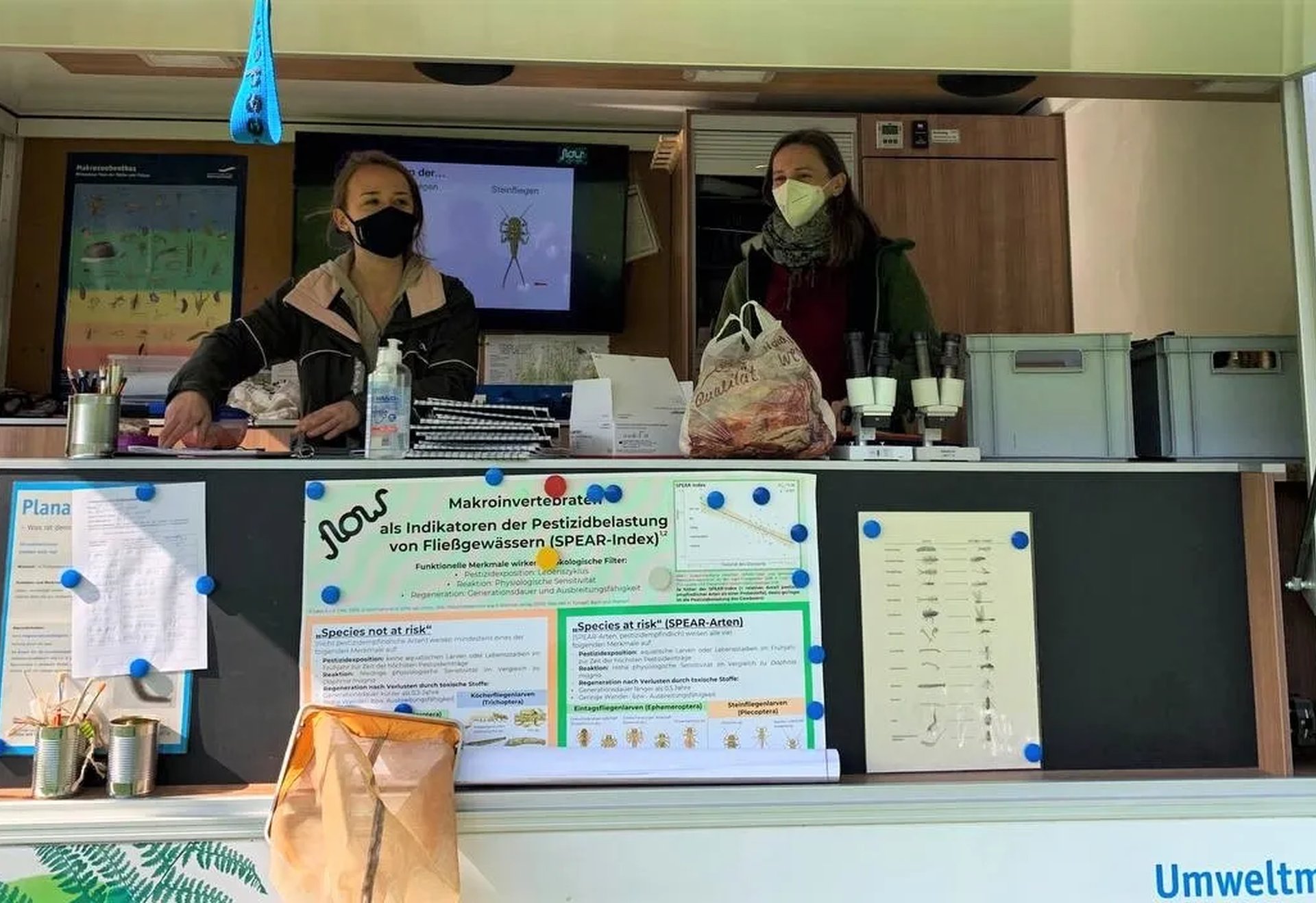 Zwei Personen mit Masken stehen in einem Umweltmobil hinter einem Tresen mit Infoplakaten und Materialien zur Umweltbildung.