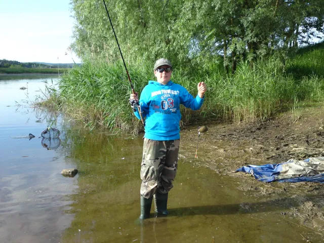 Eine Person steht mit Angelrute am Ufer eines Sees, trägt Outdoor-Kleidung und Gummistiefel.