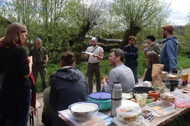 Eine Gruppe von Menschen sitzt und steht im Freien um einen Tisch mit Essen und Getränken, im Hintergrund grüne Bäume.