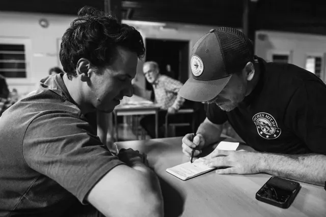 Zwei Männer sitzen an einem Tisch, einer schreibt etwas in ein Notizbuch, während der andere zuschaut. Im Hintergrund sitzen weitere Personen.