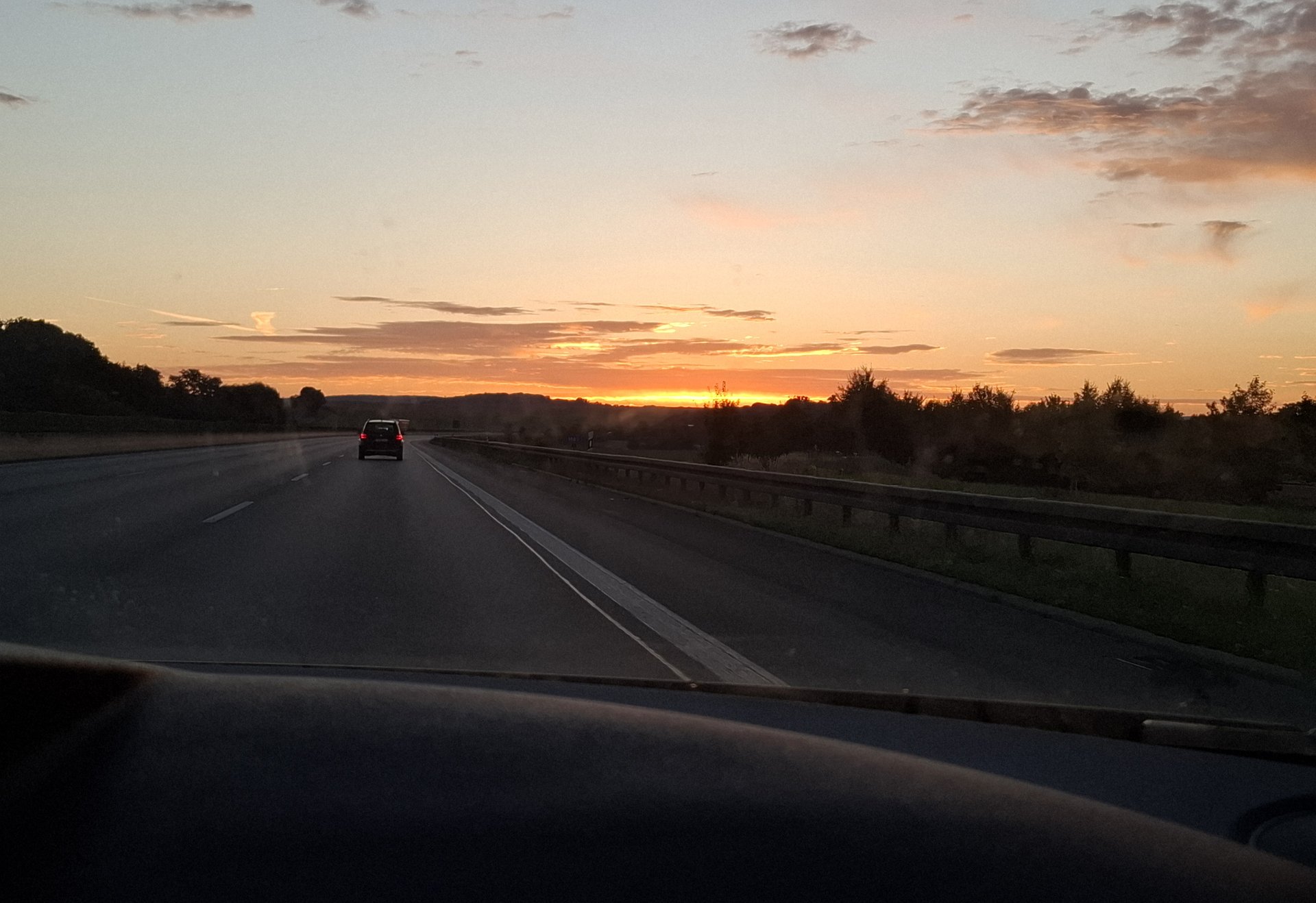 Blick aus einem Auto auf eine fast leere Autobahn bei Sonnenaufgang. Der Himmel leuchtet in warmen Orange- und Gelbtönen, während sich die Straße in die Ferne zieht.