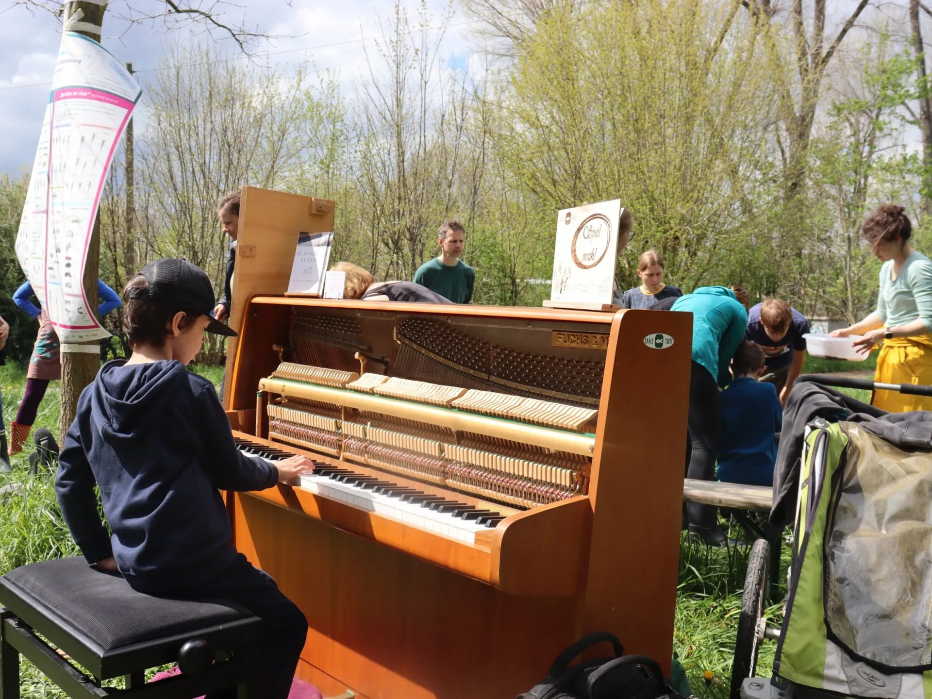 Ein Kind spielt auf einem offenen Klavier im Freien, umgeben von mehreren Menschen in einer grünen, natürlichen Umgebung.