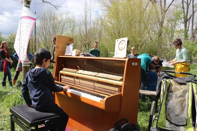 Ein Kind spielt auf einem offenen Klavier im Freien, umgeben von mehreren Menschen in einer grünen, natürlichen Umgebung.