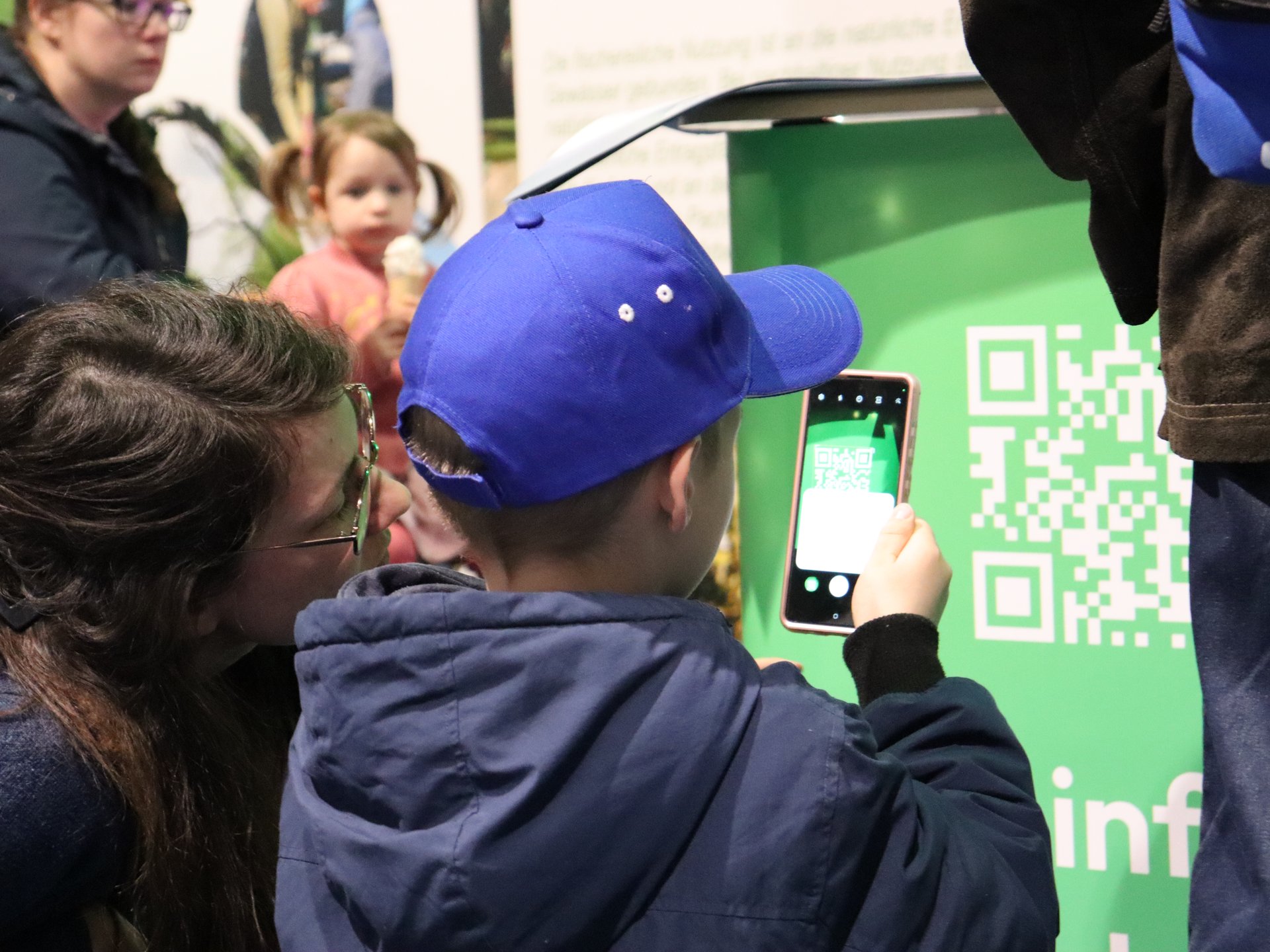 Ein Kind scannt mit einem Smartphone einen großen QR-Code auf einem grünen Schild, eine Frau schaut ihm dabei zu.