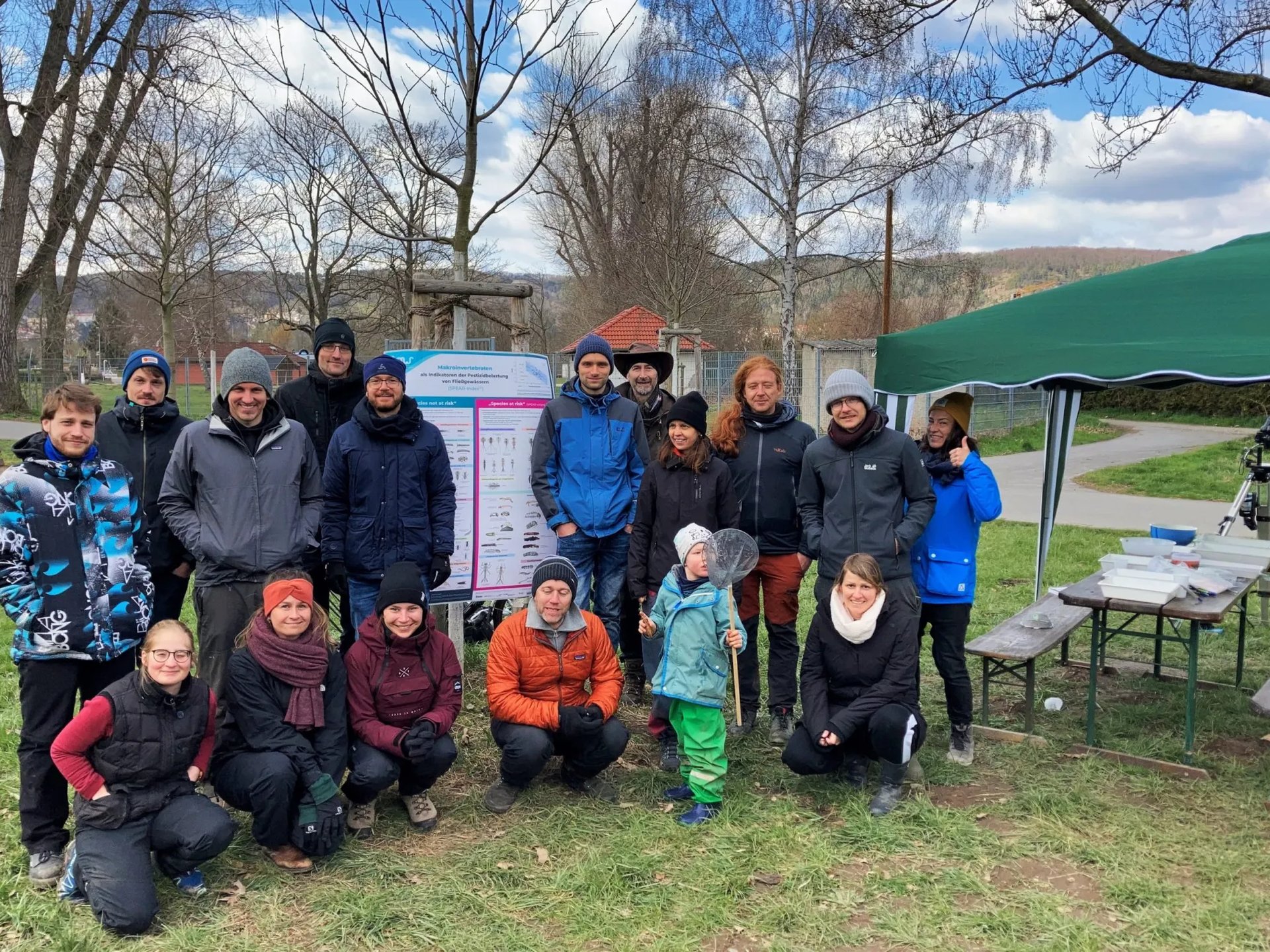 Eine Gruppe von Menschen steht und hockt im Freien vor einem Infoposter, neben ihnen ein grünes Pavillon und Tische mit Material; es ist kühl, alle tragen warme Kleidung.