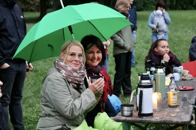 Zwei Frauen sitzen an einem Holztisch im Freien, eine hält einen grünen Regenschirm, während andere Menschen im Hintergrund stehen oder sitzen. Auf dem Tisch stehen Thermoskannen, Getränke und eine Kerze.