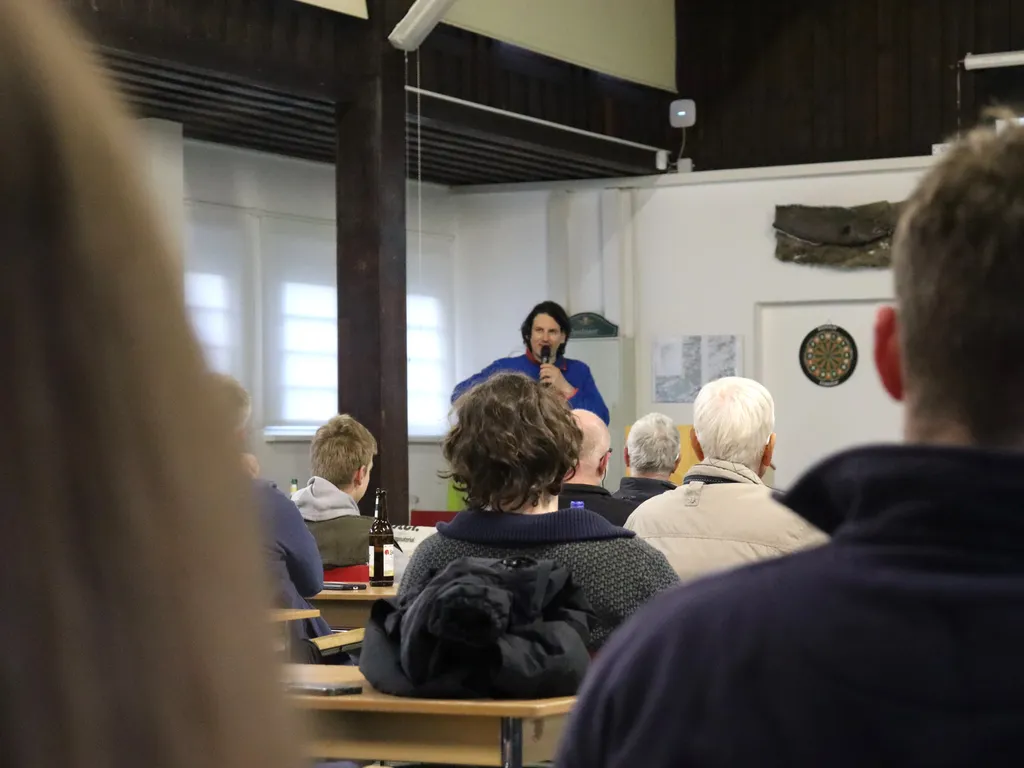Eine Person steht vorne in einem Raum und spricht vor mehreren sitzenden Zuhörerinnen und Zuhörern.