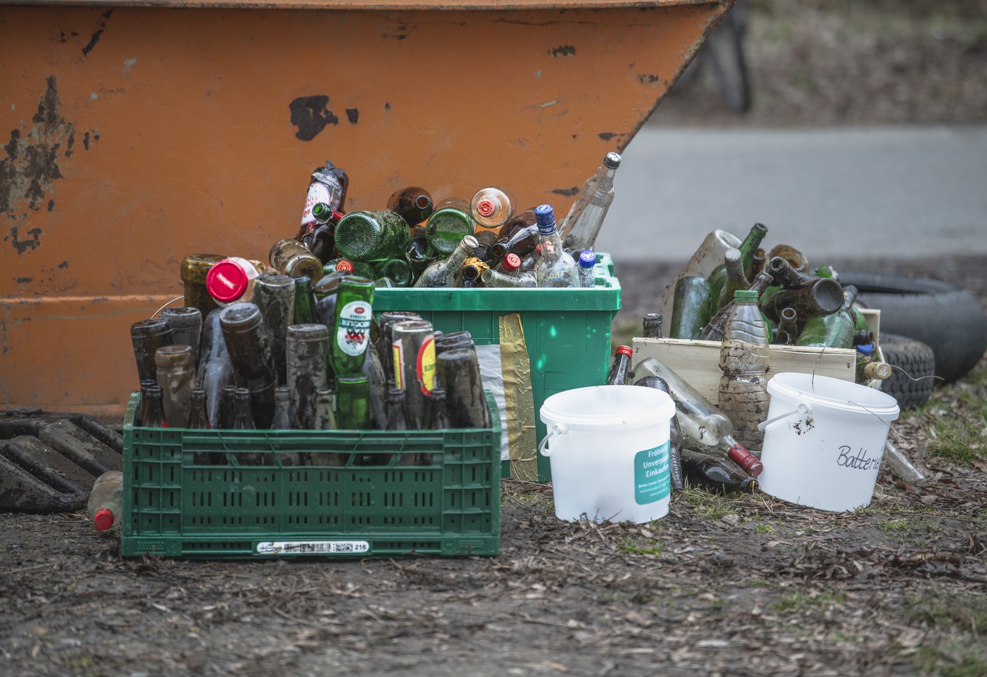 Mehrere Kisten und Eimer, gefüllt mit leeren Glasflaschen und Müll, stehen vor einem Container im Freien.