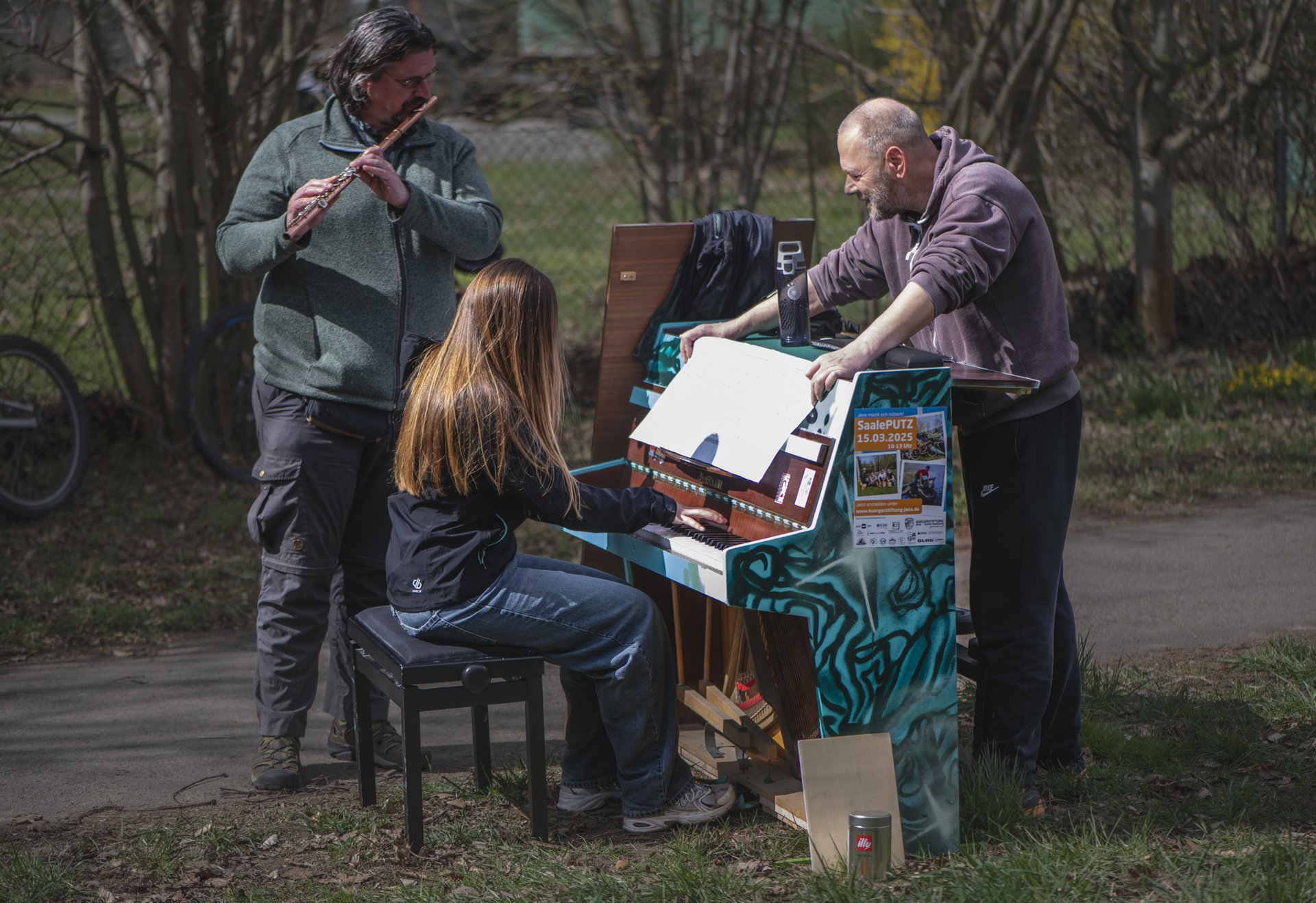 Drei Personen musizieren im Freien an einem bunt bemalten Klavier; eine Person spielt Flöte, eine sitzt am Klavier und eine weitere schaut mit Noten darauf.
