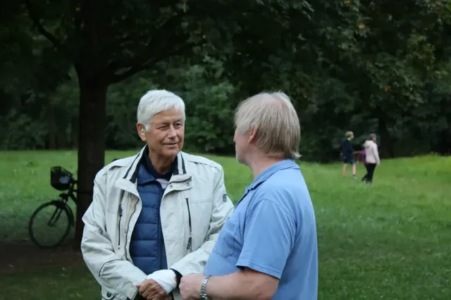 Zwei Männer unterhalten sich im Park, im Hintergrund stehen Bäume, ein Fahrrad und zwei weitere Personen.