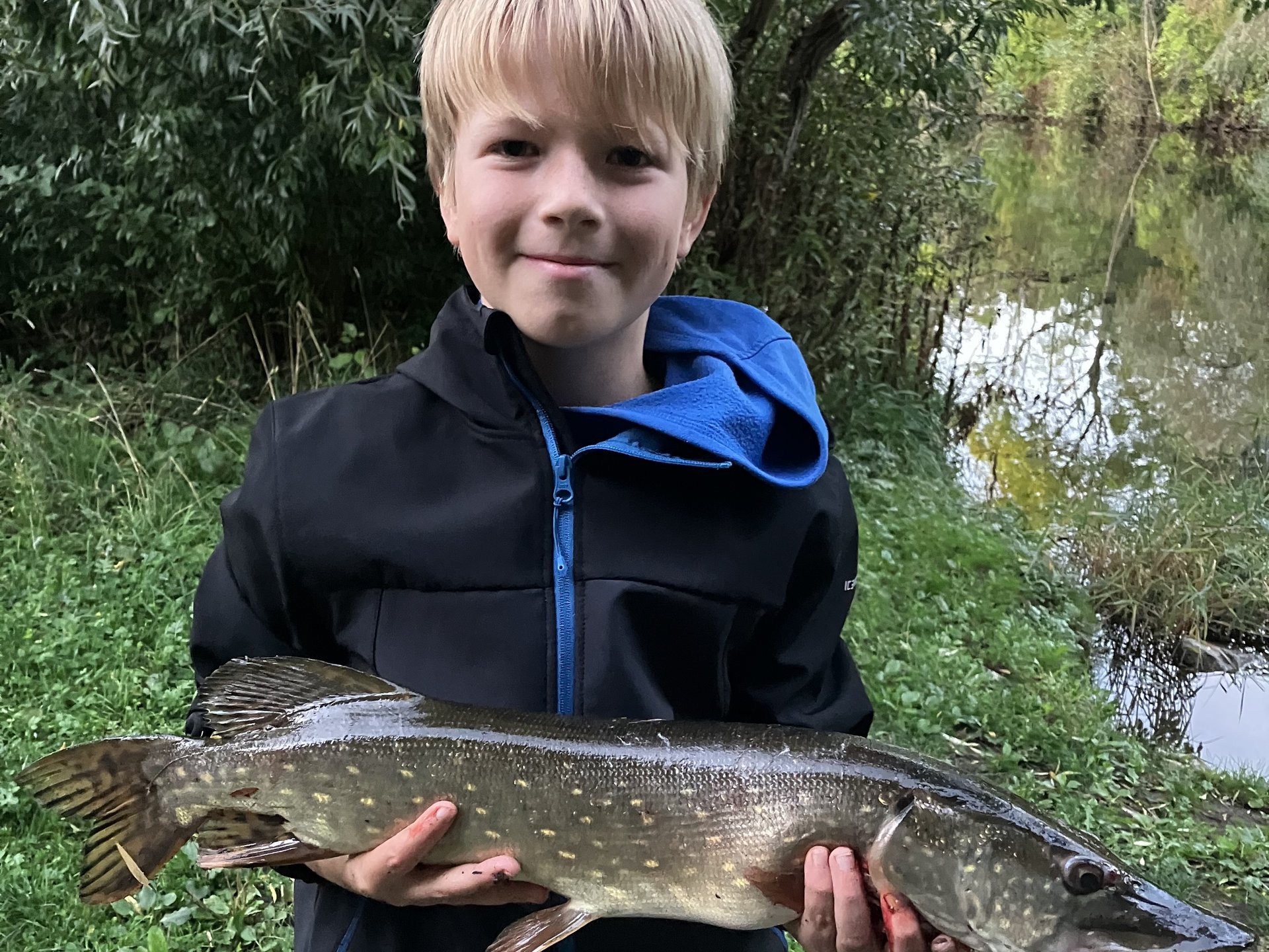 Kind mit großem Hecht in den Händen schaut in die Kamera vor Fluss