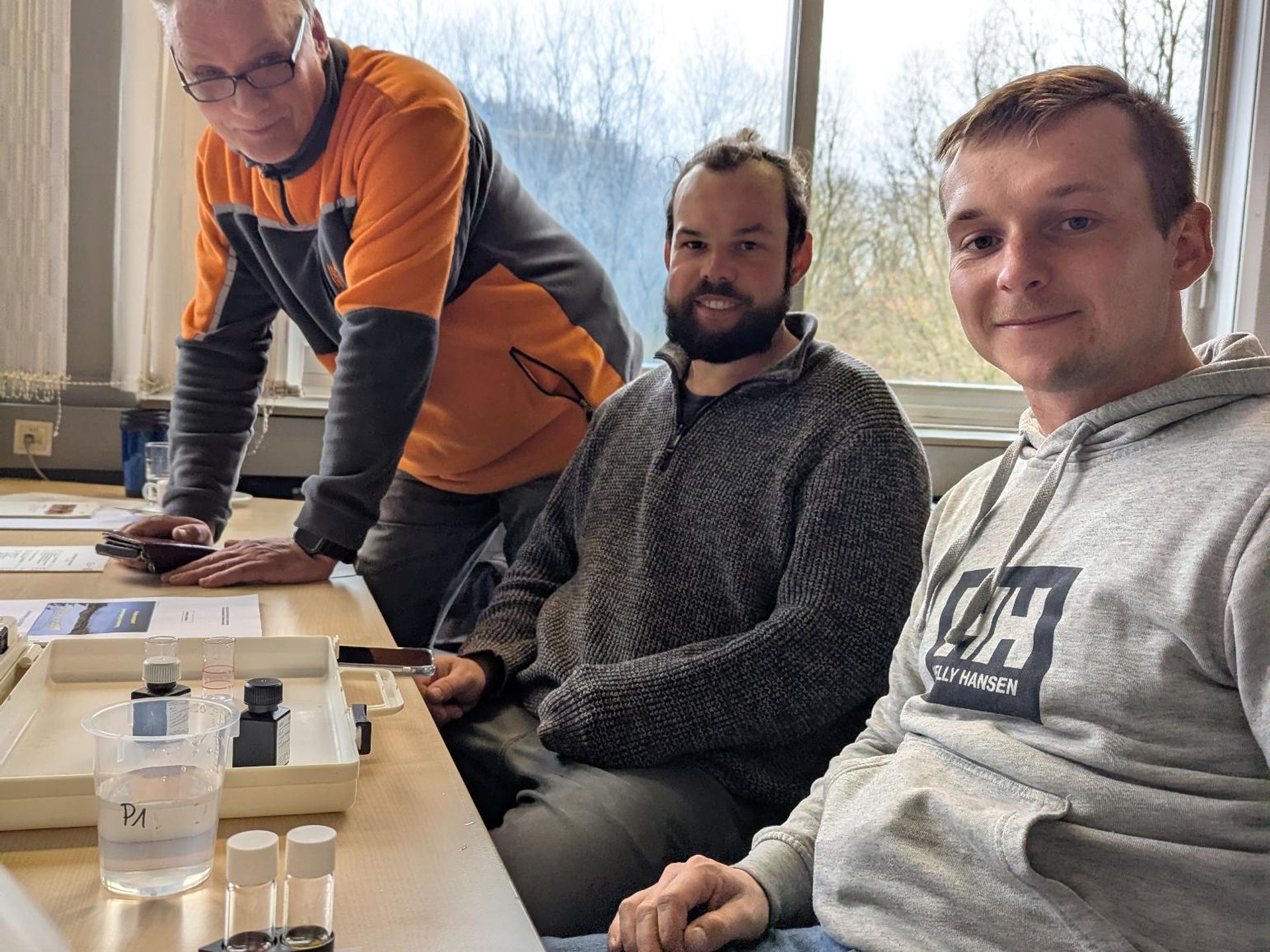 Drei Personen sitzen an einem Tisch mit Materialien zur Wasseranalyse, darunter Wasserproben, Farbkarten und kleine Messbehälter. Im Hintergrund sieht man ein großes Fenster mit Blick auf Bäume.