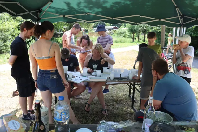 Mehrere Menschen, darunter Kinder und Erwachsene, stehen oder sitzen an einem Tisch unter einem Pavillon im Freien und beschäftigen sich mit Materialien und kleinen Bechern.