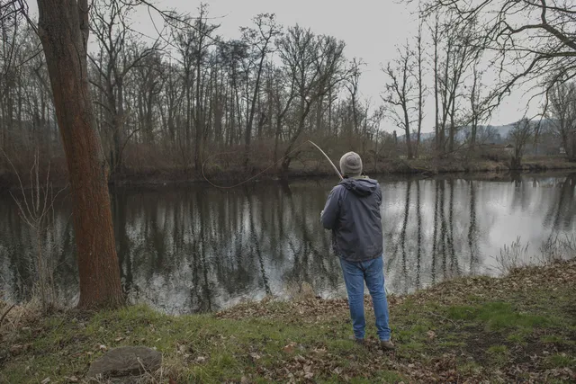 Ein Mann steht am Ufer eines Flusses und angelt, umgeben von kahlen Bäumen.