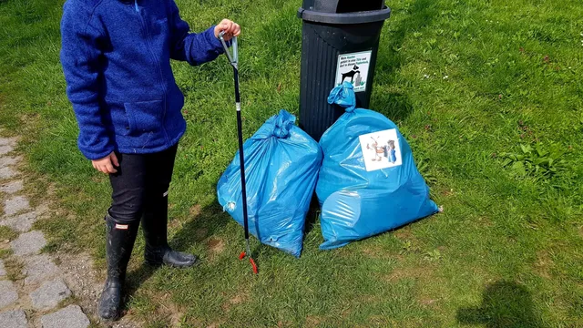 Eine Person steht neben zwei großen blauen Müllsäcken und einem Mülleimer auf einer Wiese und hält eine Greifzange.