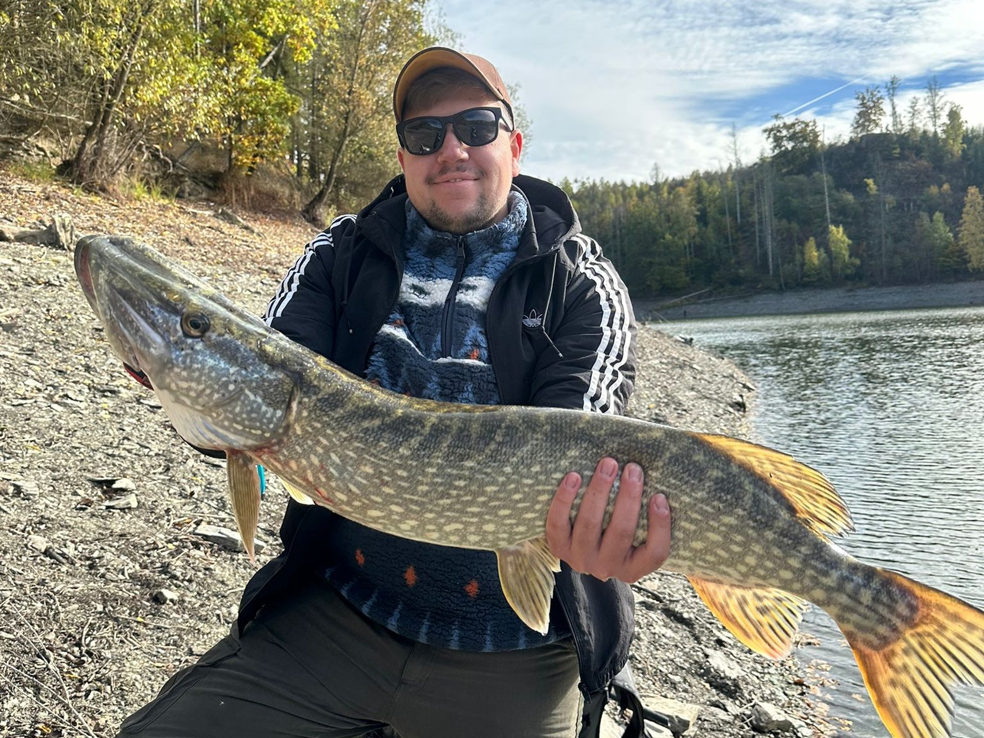 Eine Person hält einen großen Hecht vor einem felsigen Seeufer mit herbstlicher Vegetation.