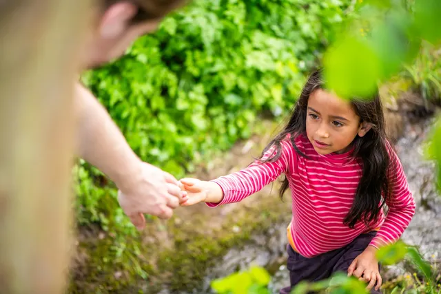 Ein Mädchen in einem gestreiften Pullover greift nach der Hand einer anderen Person, um beim Überqueren eines kleinen Bachs zu Hilfe zu bekommen.