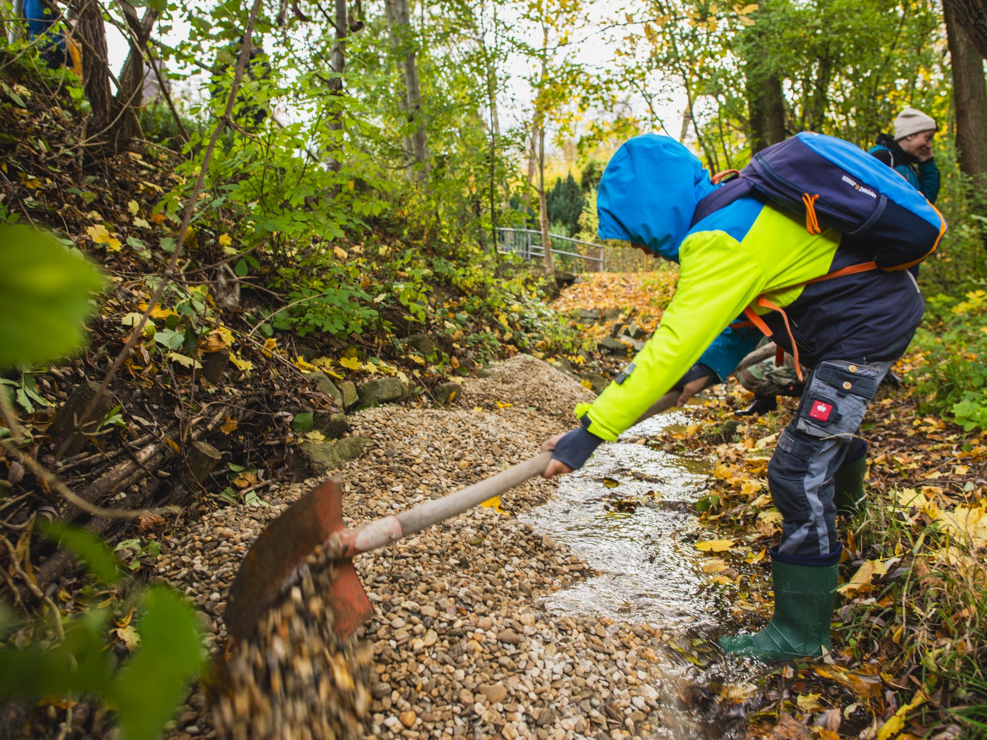 Ein Jugendlicher in wetterfester Kleidung schaufelt Kies in einen kleinen Bach, um neue Lebensräume für Fische zu schaffen.