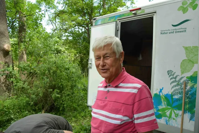 Ein älterer Mann mit grauen Haaren steht lächelnd vor einem Natur-Infomobil in einer grünen, bewaldeten Umgebung.