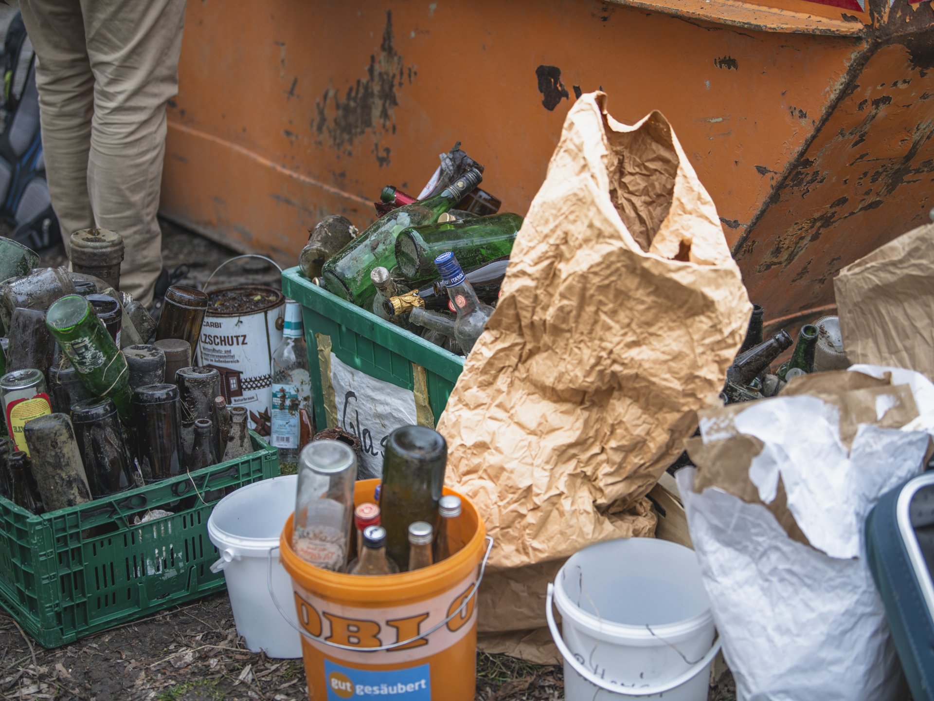 Mehrere Kisten und Eimer voller leerer und schmutziger Glasflaschen sowie Papiertüten stehen vor einem orangefarbenen Container im Freien.