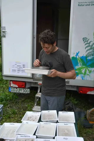 Ein Mann untersucht draußen vor einem Fahrzeug mit der Aufschrift "Natur und Umwelt" eine weiße Wanne, vor ihm stehen mehrere mit Erde und Proben gefüllte Behälter.