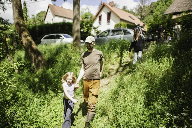 Ein Mann und ein Mädchen gehen Hand in Hand durch eine grüne, sonnige Wiese, im Hintergrund sind weitere Menschen und Häuser zu sehen.