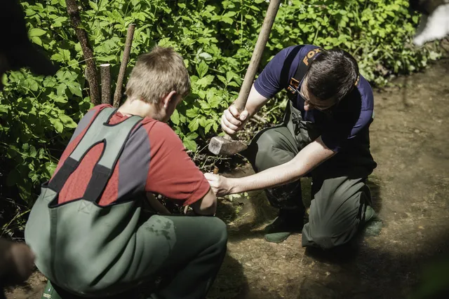 Zwei Personen arbeiten kniend mit Werkzeugen an einem Bachlauf im Grünen.
