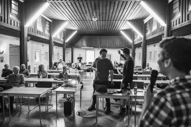 In einem großen Saal sitzen mehrere Menschen an Tischen, während vorne drei Personen stehen, von denen eine ein Mikrofon hält und spricht.