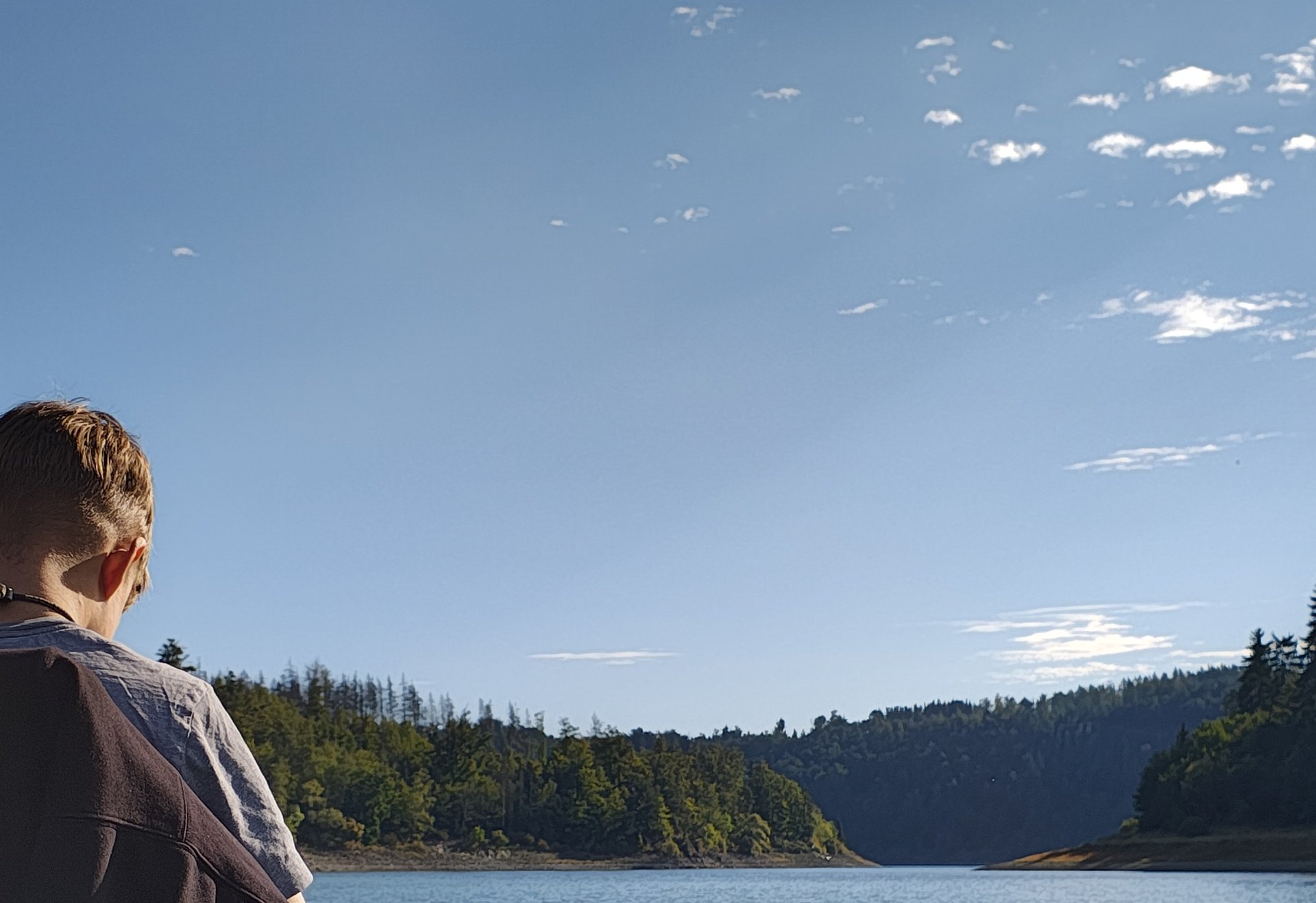 Ein junger Angler sitzt am Heck eines Bootes auf einem See, umgeben von bewaldeten Hängen und blauem Himmel mit lockeren Wolken.