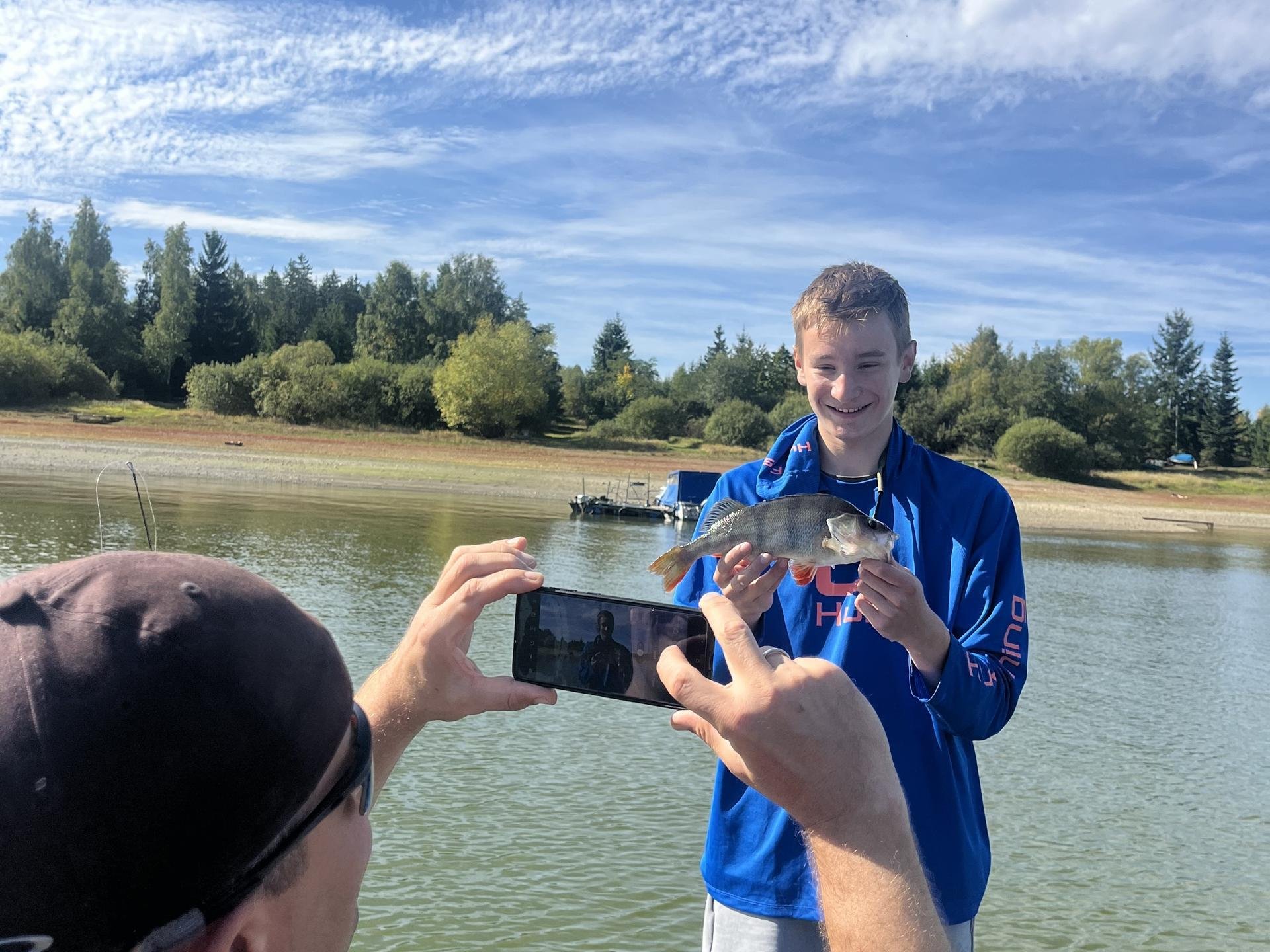 Ein junger Mann steht auf einem Boot und hält einen Fisch in den Händen, während eine andere Person ein Foto von ihm macht. Im Hintergrund sind Bäume und ein Seeufer zu sehen.
