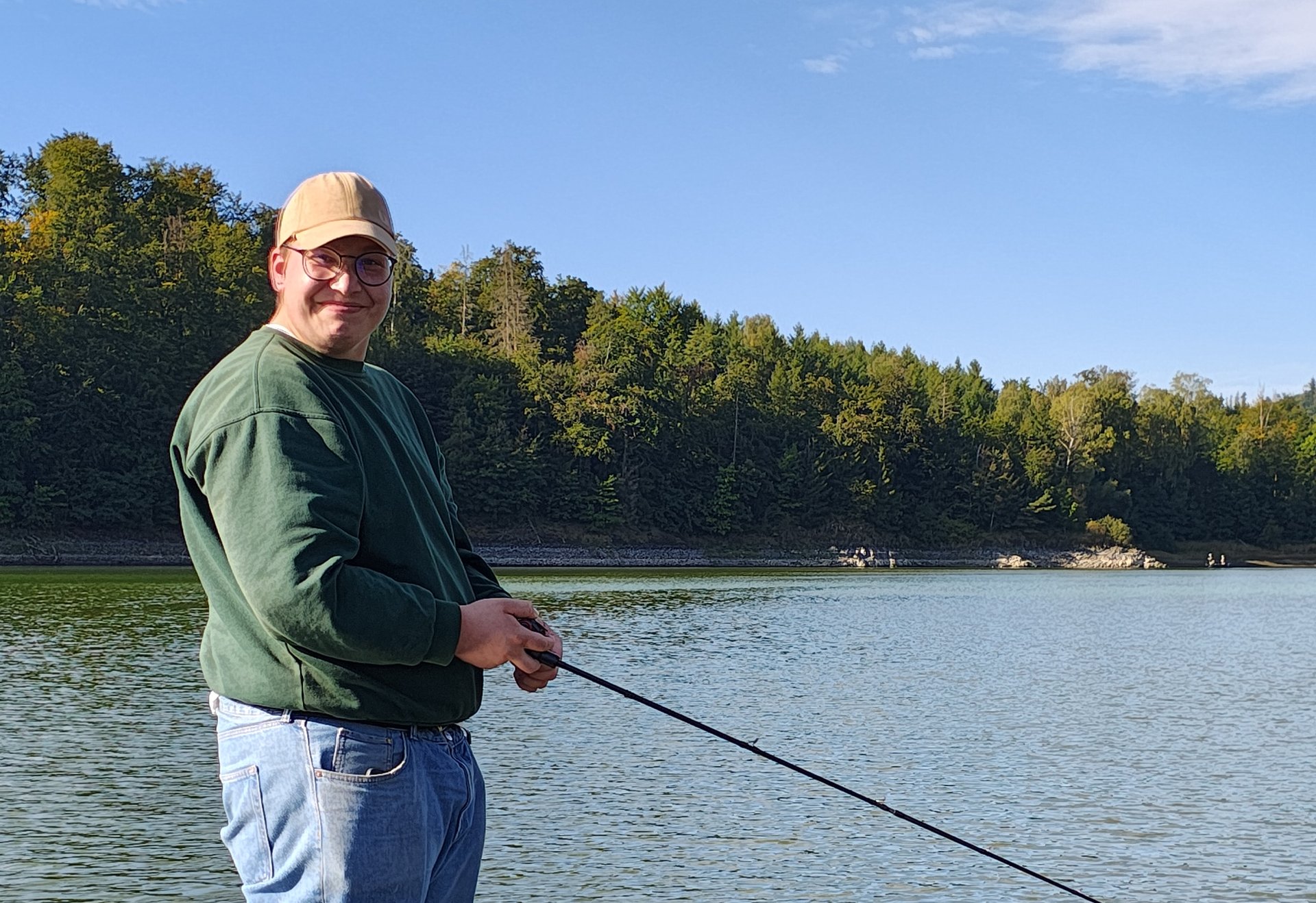 Ein Angler steht auf einem Boot, hält die Angelrute über das Wasser und blickt auf den sonnigen See.