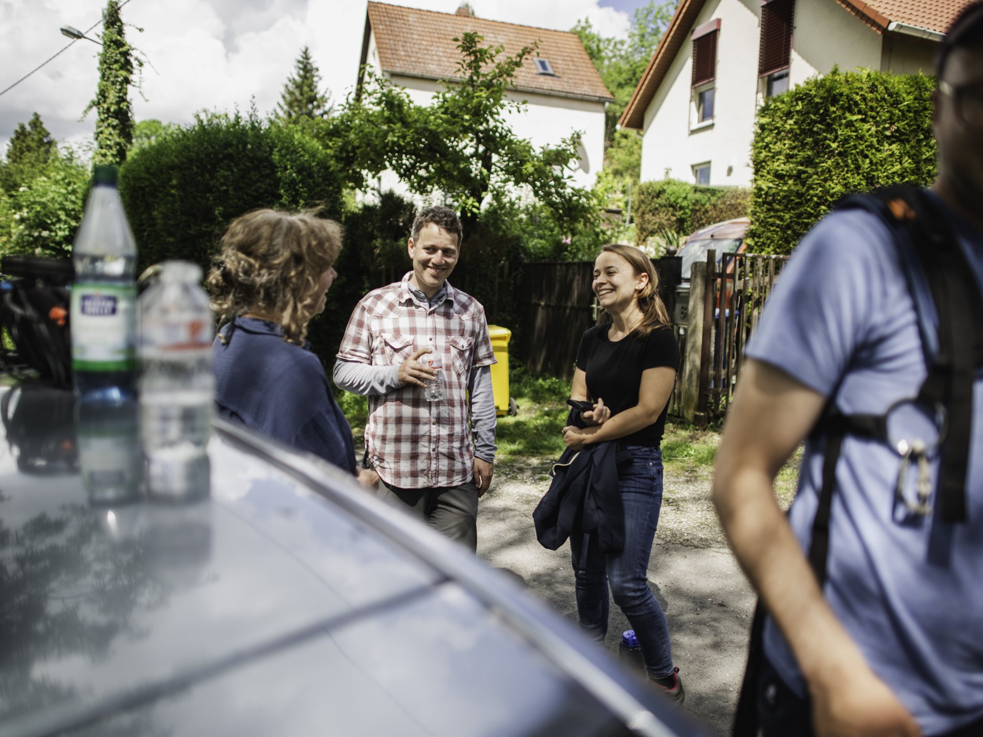 Mehrere Personen unterhalten sich im Freien vor Häusern, im Vordergrund stehen Wasserflaschen auf einem Autodach.