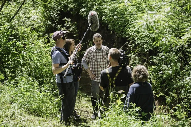 Eine Filmcrew dreht im Grünen und nimmt eine Person mit Mikrofon, Kamera und Tonangel auf.