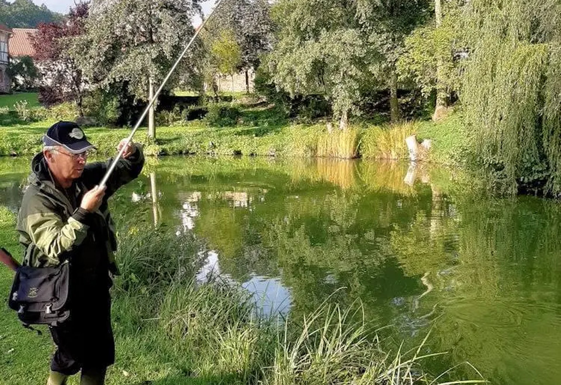 Ein Angler steht am Ufer eines grünen Teichs und hält eine Angelrute, umgeben von Bäumen und Gras.