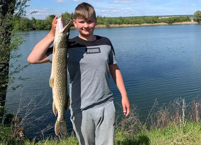 Ein Junge steht am Seeufer und hält einen großen Fisch in die Höhe.