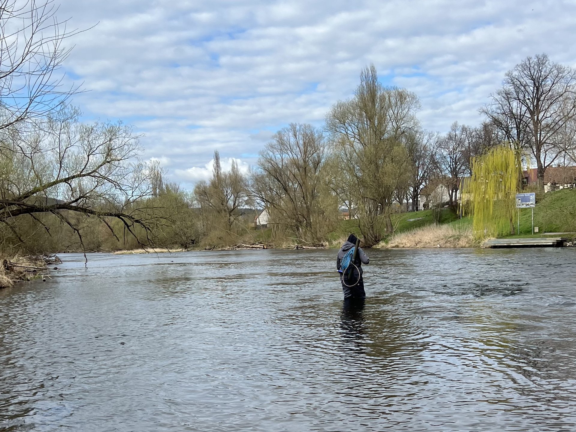 Eine Person steht in einem Fluss und angelt, umgeben von Bäumen und unter bewölktem Himmel.