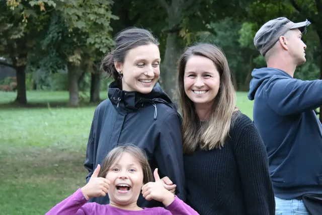 Drei lachende Personen, zwei Frauen im Hintergrund und ein fröhliches Mädchen im Vordergrund, stehen draußen im Grünen; ein Mann dreht sich im Hintergrund zur Seite.