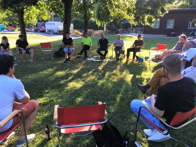 Mehrere Menschen sitzen im Kreis auf Stühlen im Freien auf einer Wiese unter Bäumen und unterhalten sich.