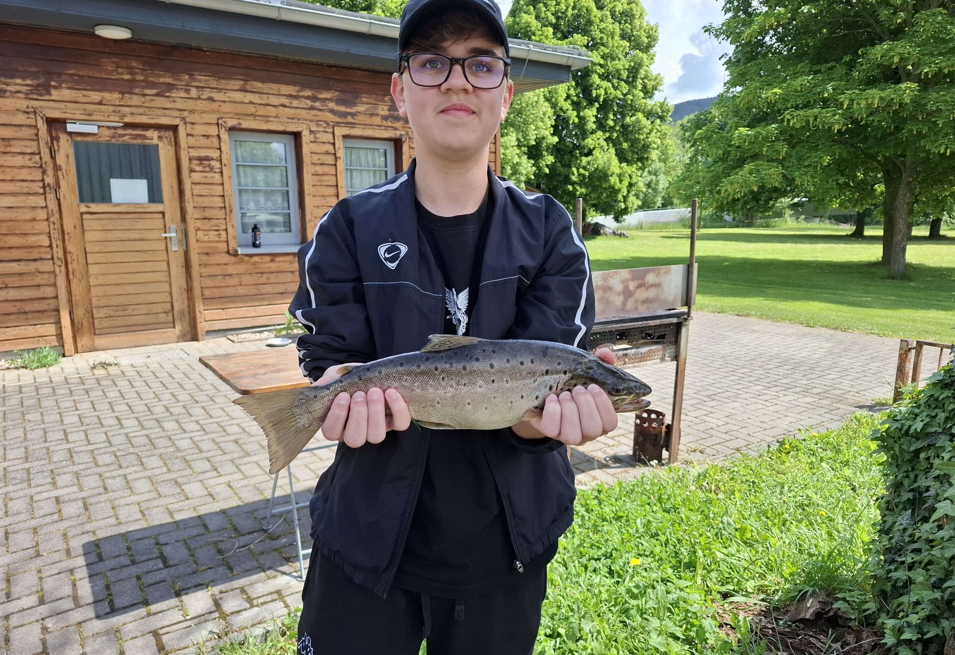 Eine Person steht im Freien und hält einen großen Fisch vor sich in den Händen. Im Hintergrund ist ein Holzhaus sowie grüne Bäume und Wiese zu sehen.