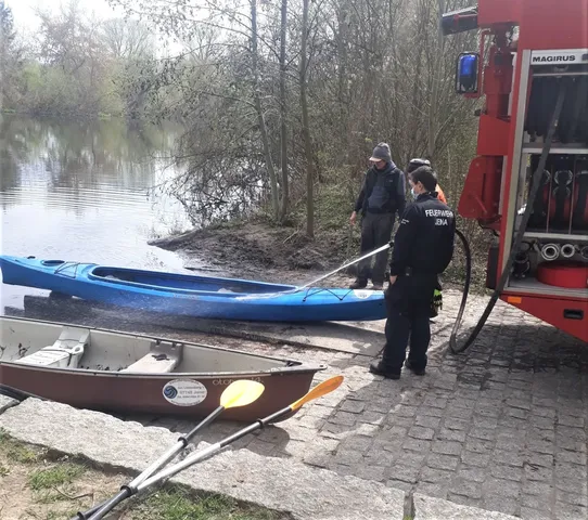 Zwei Personen der Feuerwehr reinigen ein blaues Kajak mit einem Wasserschlauch neben einem Feuerwehrfahrzeug am Ufer eines Sees.