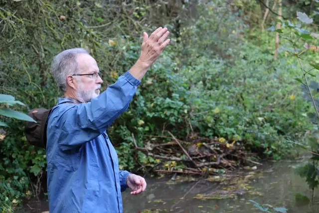 Ein älterer Mann mit grauem Bart steht am Ufer eines kleinen Bachs im Wald und hebt eine Hand, als würde er etwas erklären oder zeigen.